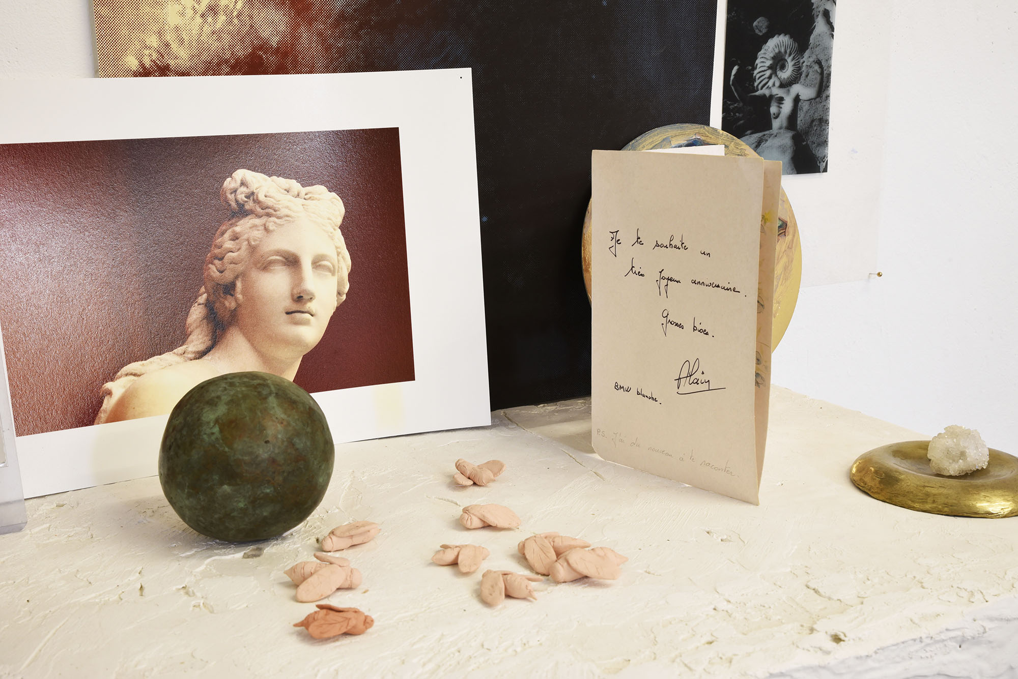 Gros plan sur les choses posées sur la cheminée : des abeilles en céramique rose pâle, une boule en métal, un cristal posé sur une soucupe en métal doré, une carte d'anniversaire signée par Alain, une photo d'une sculpture d'Aphrodite sur fond rouge et une photo noir et blanc d'une femme a tête de coquillage.